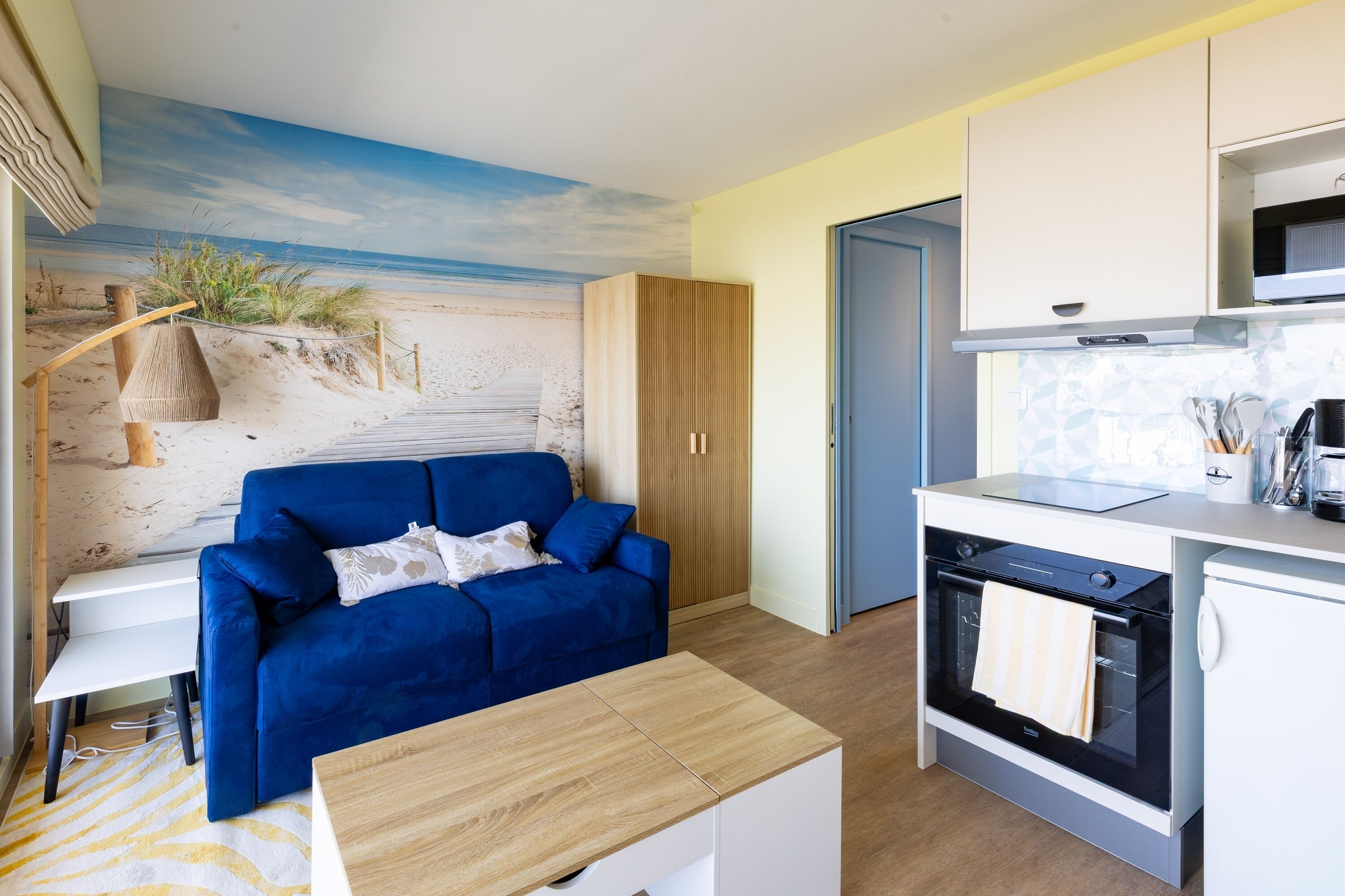 Un espace de vie compact agrémenté d’une fresque murale représentant une plage, d’un canapé bleu, d’une kitchenette et d’une table en bois.