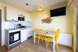 A compact kitchen with a microwave, stove, and sink, a dining table with yellow chairs, and a flat-screen TV mounted on the wall. - Ker Mor - À 150 m de la plage (Quiberon)