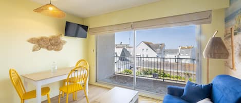 Un salon moderne comprenant un canapé, une table à manger avec des chaises, un téléviseur fixé au mur et une grande fenêtre donnant sur le quartier.