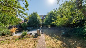 Ein Weg, der durch einen Garten mit verschiedenen Pflanzen und Bäumen führt, eine Bank und ein Gebäude im Hintergrund.