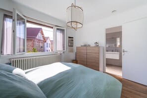 Une chambre à coucher comprenant un lit, une commode, une fenêtre avec des rideaux, un luminaire suspendu et une vue sur une maison à l’extérieur.