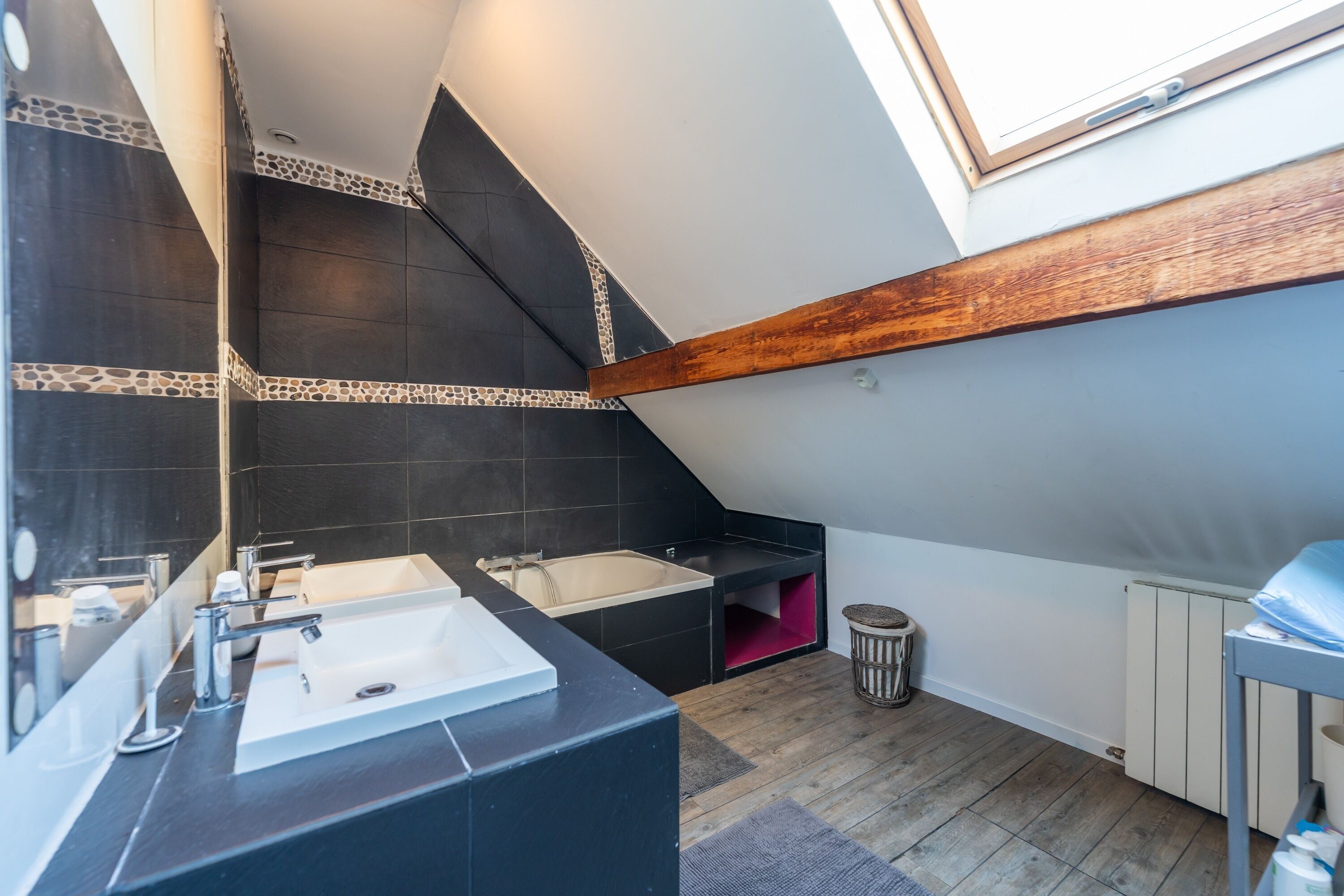 A modern bathroom with a skylight, dark tiles, a wooden beam, and a small trash bin.