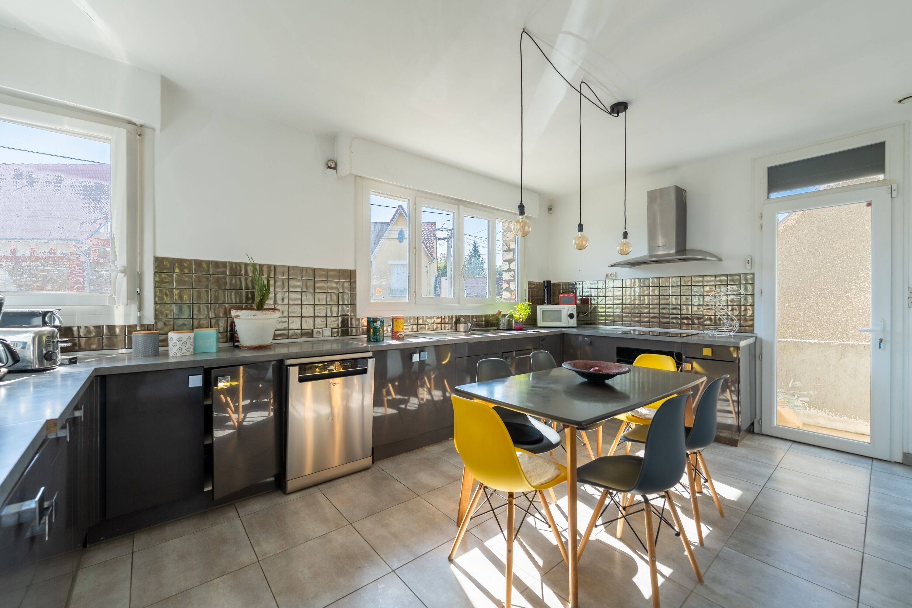 A modern kitchen with a central island, a dining table with chairs, and a view of the outside through large windows.