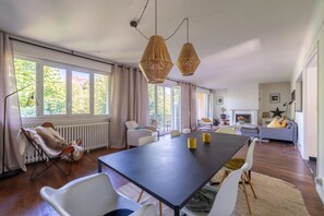 A modern living room with a large dining table, white chairs, and a sofa. There are large windows with curtains, a fireplace, and a star decoration on the wall. - La Maison Rabier - Gare La Barre Ormesson à 300 m (Deuil-la-Barre)