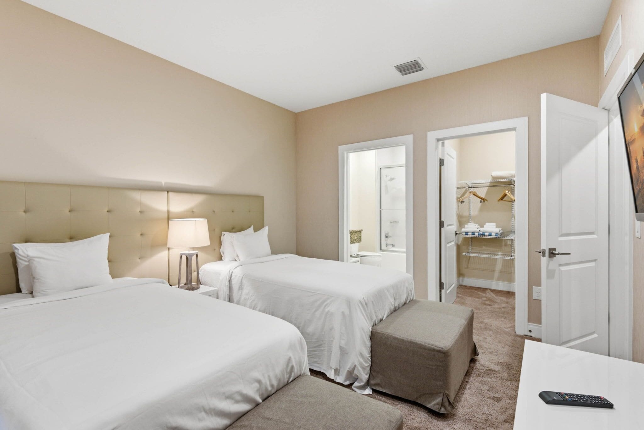 Une chambre d’hôtel avec deux lits, une table de chevet, une lampe, une salle de bain visible à travers une porte ouverte et une télécommande posée sur une petite table.