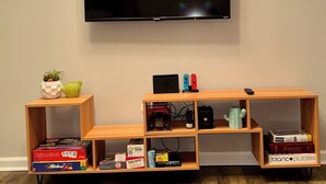 Una TV a schermo piatto montata a parete, un mobile in legno contenente giochi e libri, e una piccola pianta di cactus su uno scaffale.