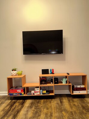 A flat-screen TV mounted on a wall, a wooden entertainment unit with games and books, and a small cactus plant on a shelf.