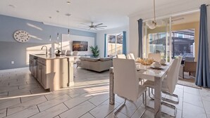 Et moderne køkken med en central ø, et spiseområde med et hvidt bord og stole, og en opholdsstue med en sofa og et fjernsyn.