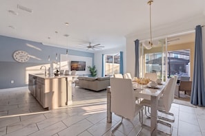 Eine moderne Küche mit einer zentralen Kochinsel, einem Essbereich mit weißem Tisch und Stühlen sowie ein Wohnbereich mit Sofa und Fernseher.