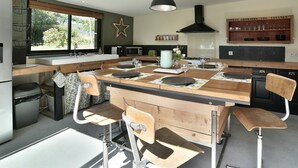 Una cocina con una isla central, sillas de madera, una lámpara colgante y un ventanal con persianas.