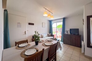 A dining area with a table set for four, chairs, a potted plant, and a television on a cabinet. - L’Évasion Claire Rive - Proche domaines viticoles (Prayssac)