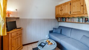 A compact living space with a grey sofa, a small round table with a tray, a television on a wooden cabinet, and a bookshelf with books. - Le Petit Jardin du Criou - Station de ski à 800 m (Samoëns)