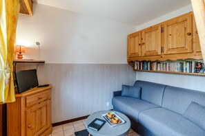 A compact living space with a grey sofa, a small round table with a tray, a television on a wooden cabinet, and a bookshelf with books. - Le Petit Jardin du Criou - Station de ski à 800 m (Samoëns)