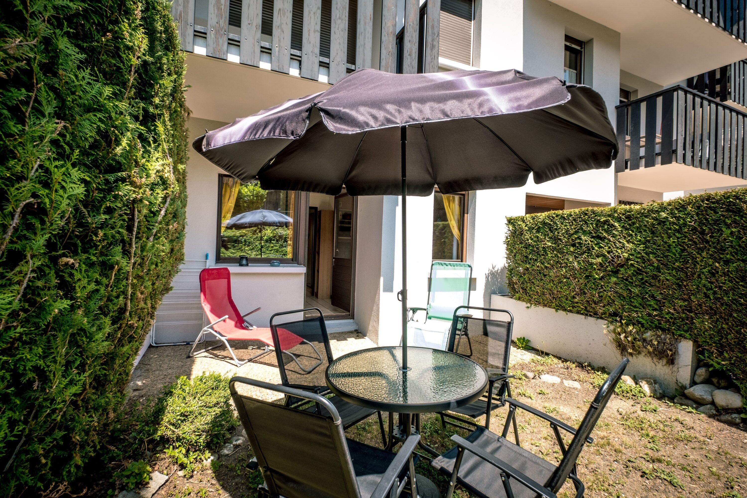 An outdoor patio with a round table and chairs, a red chair, and a large umbrella providing shade.