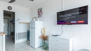 A modern living room with a flat-screen TV mounted on the wall, a white refrigerator, a small table with a vase, and a fan. - Le Flamant Rose - Climatisé et à 200 m de la plage (Mauguio)