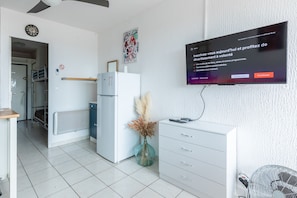 A modern living room with a flat-screen TV mounted on the wall, a white refrigerator, a small table with a vase, and a fan. - Le Flamant Rose - Climatisé et à 200 m de la plage (Mauguio)