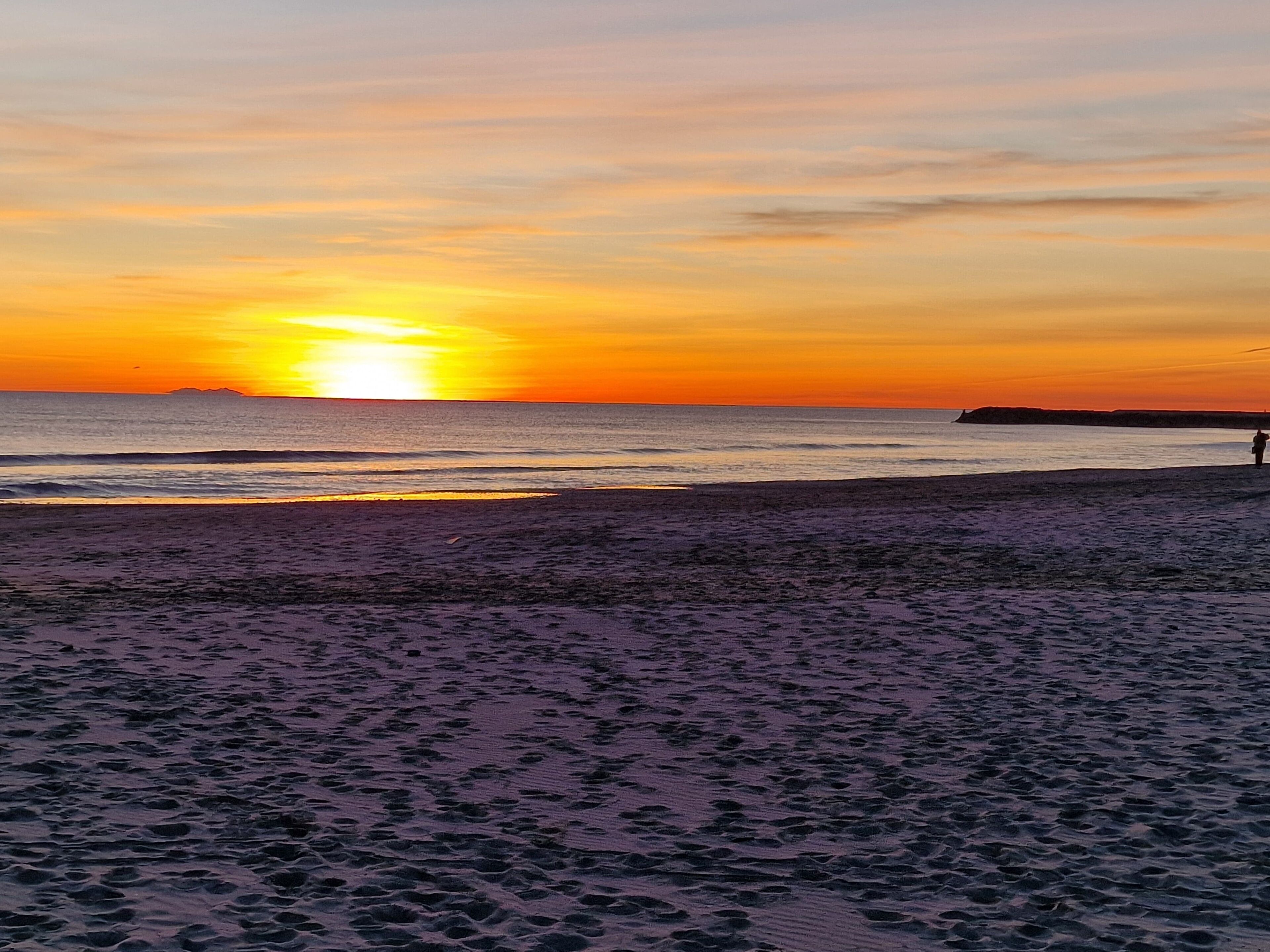Ein Strand mit Fußspuren, die zum Wasser führen, eine Person am Ufer und ein Sonnenuntergang im Hintergrund.