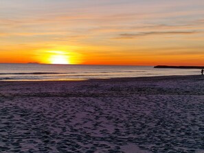 Ranta, jossa näkyy jalanjälkiä, jotka johtavat veteen, henkilö seisoo rannan reunalla ja taustalla auringonlasku.