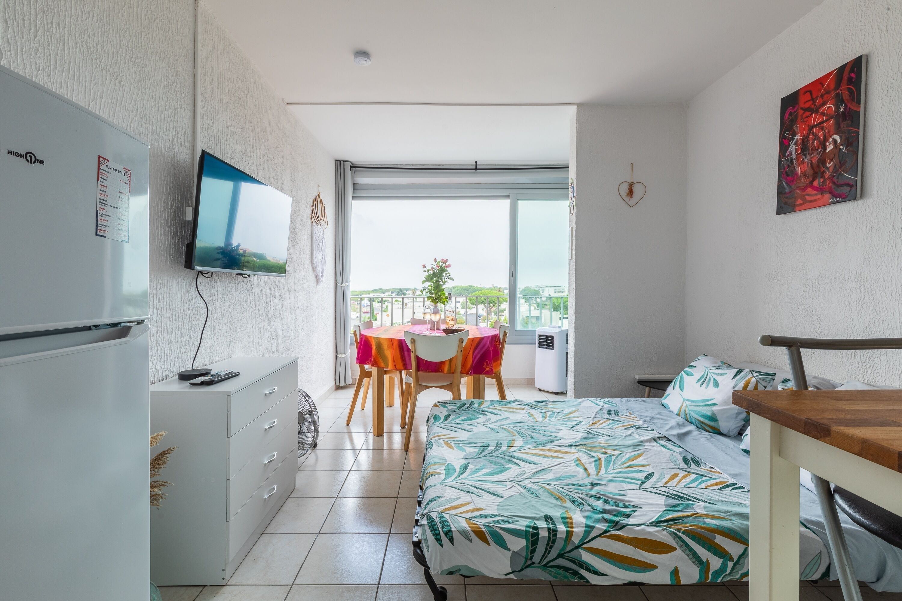 A compact hotel room with a bed, a desk, a dining table, and a balcony with a view.