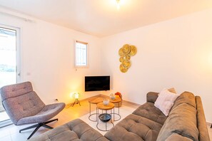 Ein modernes Wohnzimmer mit grauem Sofa, einem gemusterten Sessel, einem kleinen Holztisch und einem flachbildfernseher an der Wand.