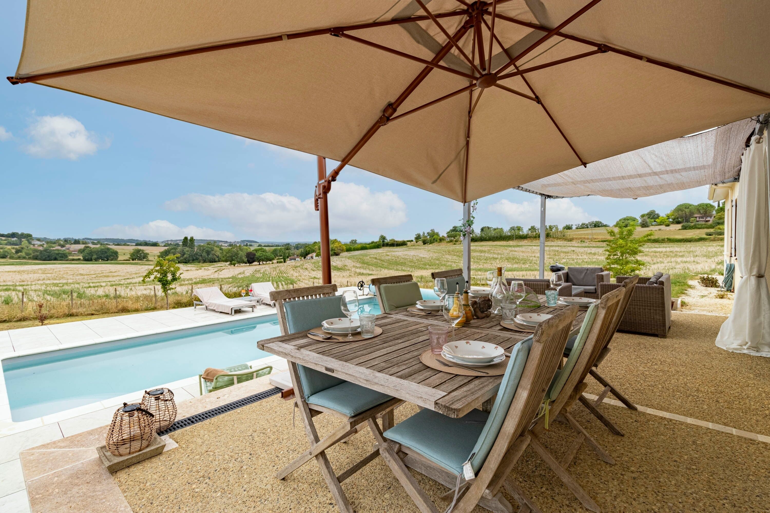 Un área de comedor al aire libre con piscina, muebles de madera y vistas al campo.
