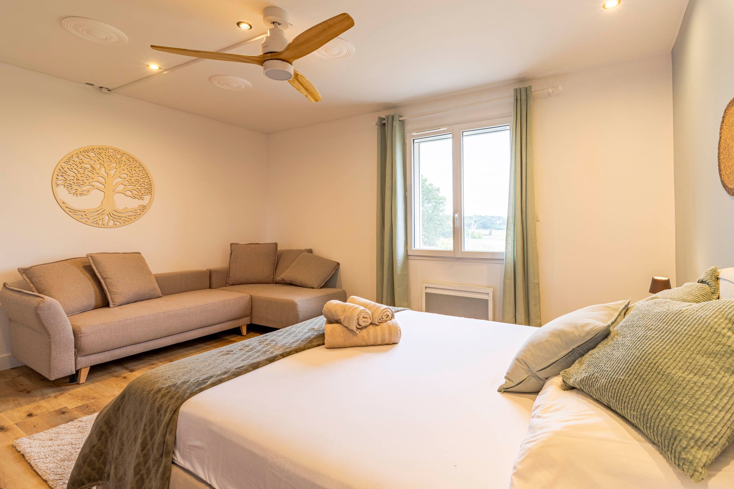 A bedroom with a bed, a sofa, a ceiling fan, a window with curtains, and a wall decoration.