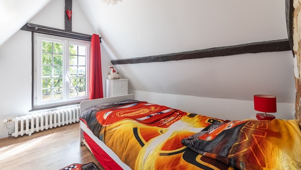 A room with a sloped ceiling, a bed with a colorful bedspread, a window with a view of greenery, a radiator, and a red lamp on a bedside table. - La Maison Maudanaur - Maison familiale avec jardin (Morainville-Jouveaux)