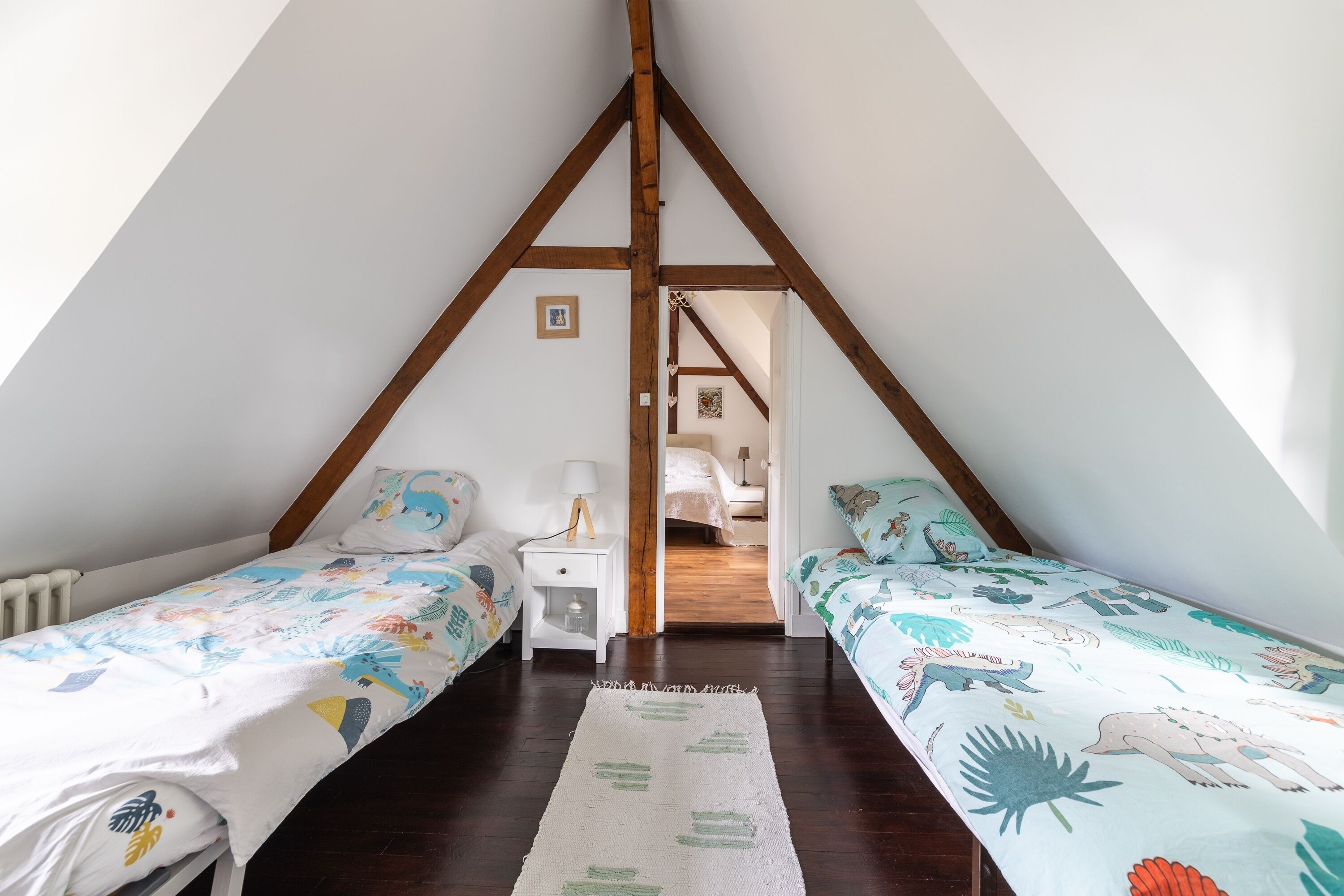 A bedroom with two single beds, a white nightstand with a lamp, and a wooden ceiling structure.