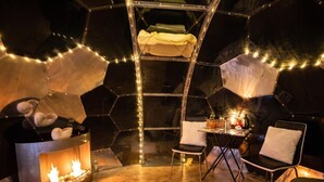 Ein Zimmer mit einer sechseckigen Wandgestaltung, einer zentralen Sitzecke mit Sesseln, einem kleinen Tisch und einem Kamin mit sichtbaren Flammen.