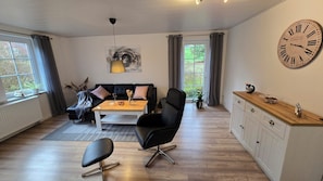 A modern living room with a sofa, coffee table, and a dining area with chairs. A large wall clock is visible. - Cozy vacation rental in the picturesque town of Uehrde (Osterode am Harz)