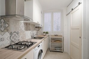 A compact kitchen with a gas stove, a washing machine, and a small window with patterned curtains. - GuestHost - Azurea Home: Lovely & Spacious Apartment for 6 (Sanremo)