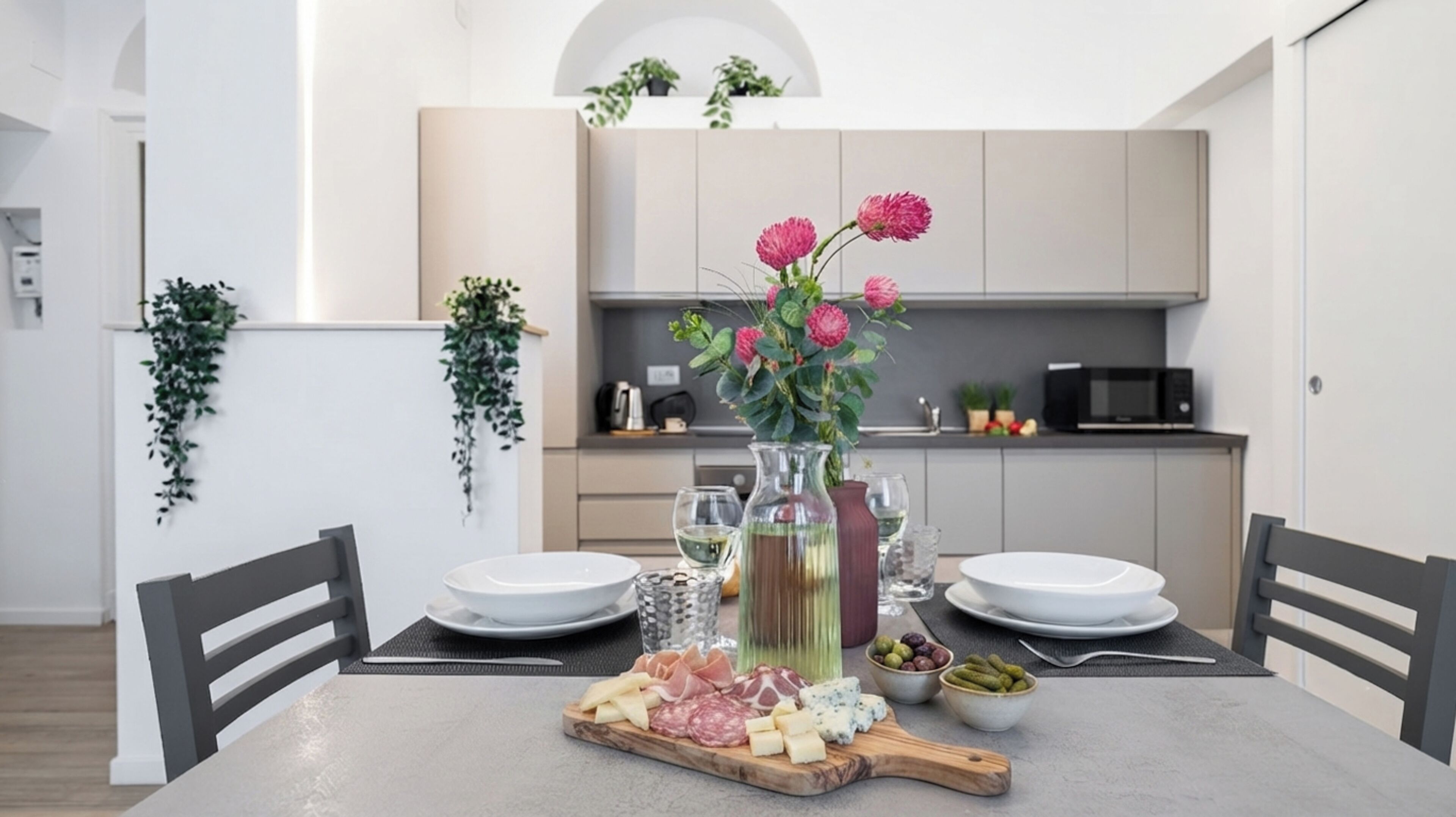 A dining table set with a variety of cheeses, cured meats, and olives, accompanied by a bottle of wine and a vase with flowers.