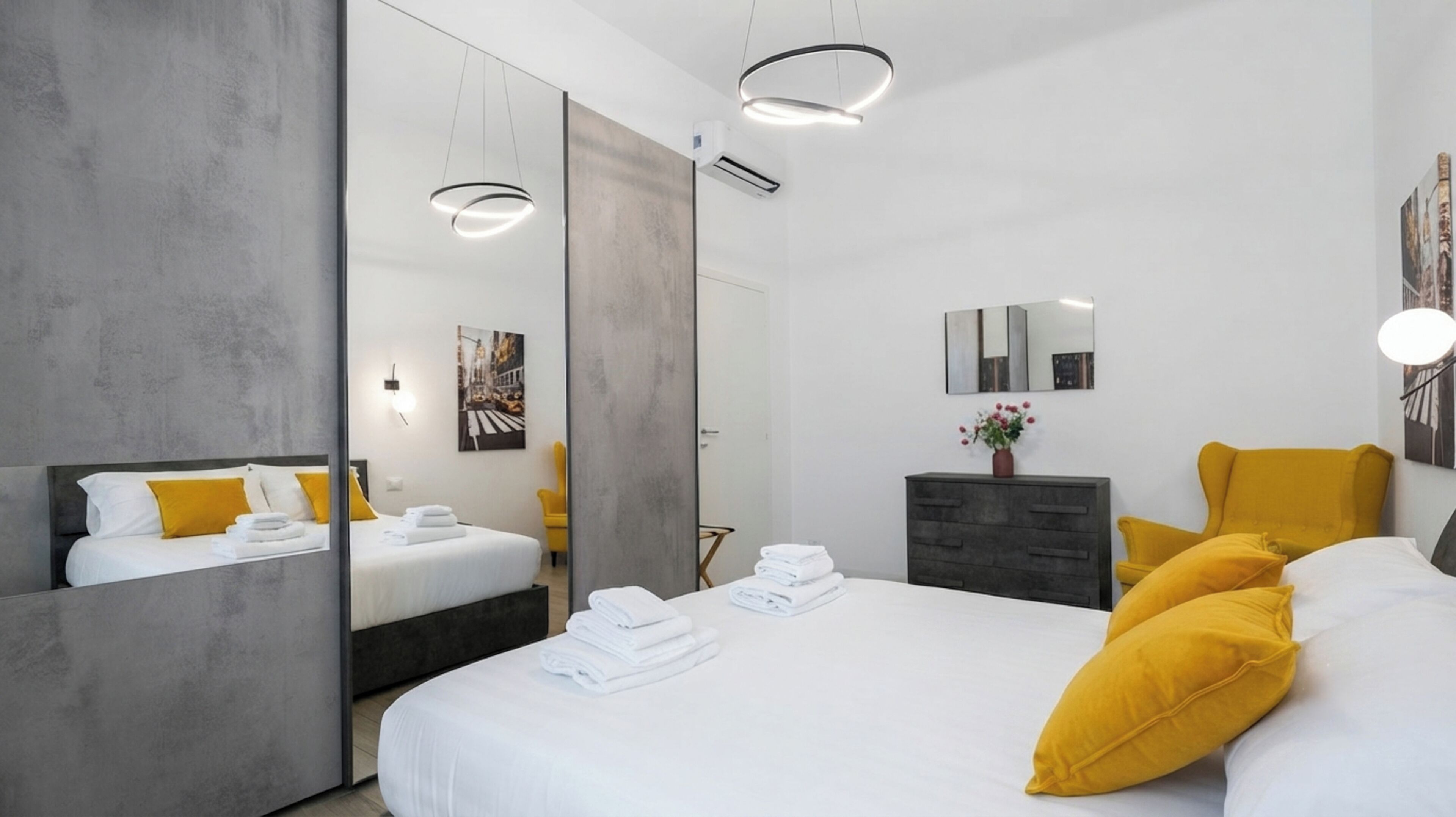 A modern hotel room with a large bed, a wardrobe, a mirror, and a small table with towels.