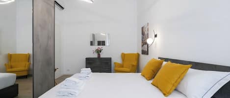 A modern hotel room with a large bed, two chairs, a dresser, a mirror, and a wall-mounted light fixture.