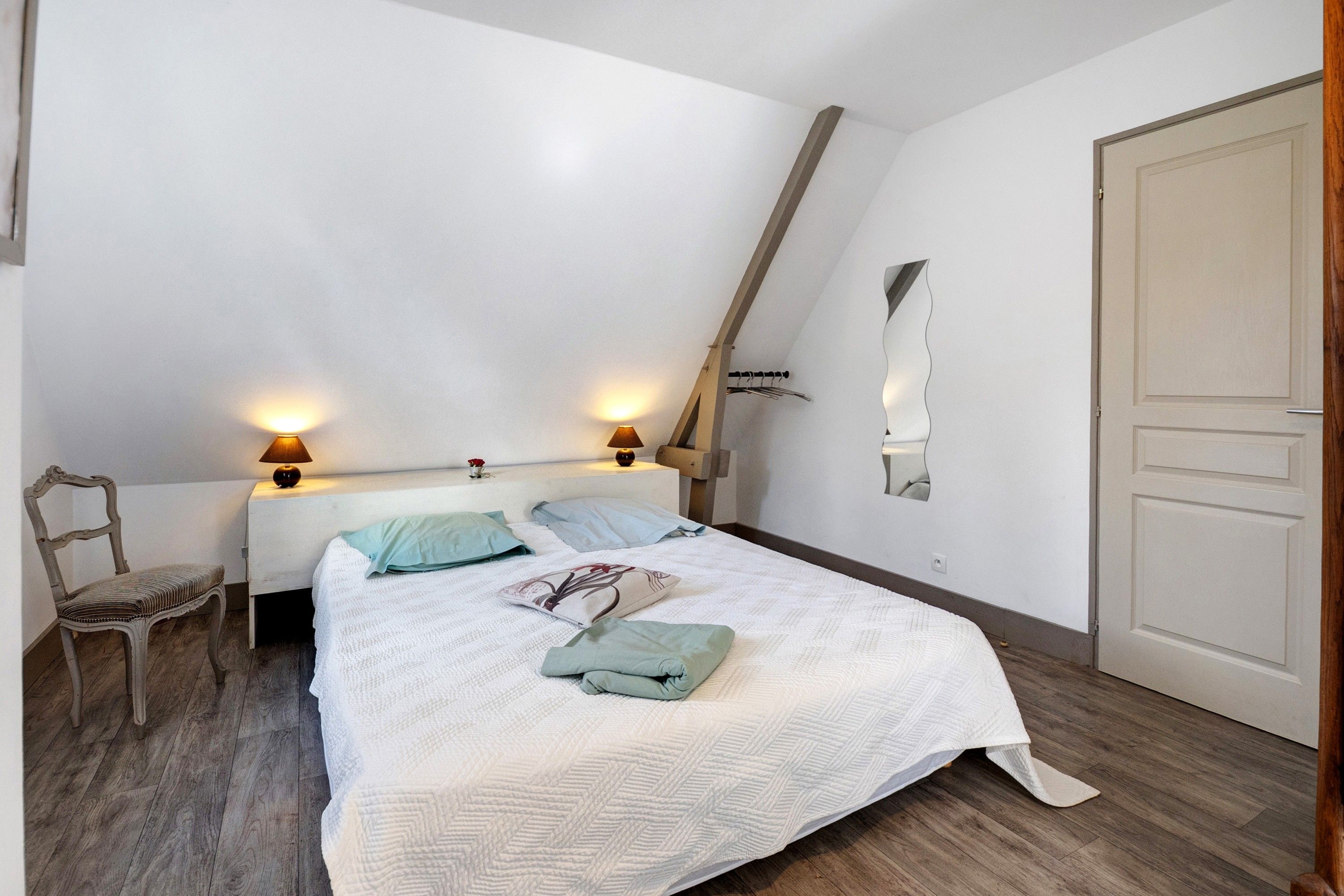 A bedroom with a bed, two bedside lamps, a chair, a wooden floor, and a door.