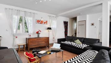 A modern living room with a black leather sofa, a wooden coffee table, a wicker chair, and a dining table.