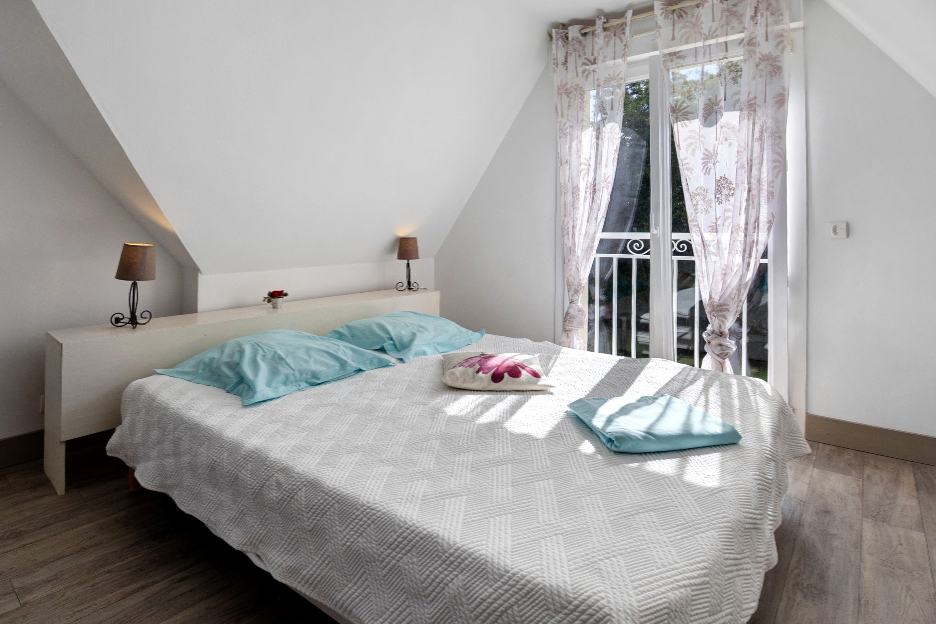 Une chambre avec un plafond en pente, un lit recouvert d’une couette blanche et agrémenté d’oreillers bleus, deux lampes de chevet et une fenêtre ornée de rideaux à motifs floraux.