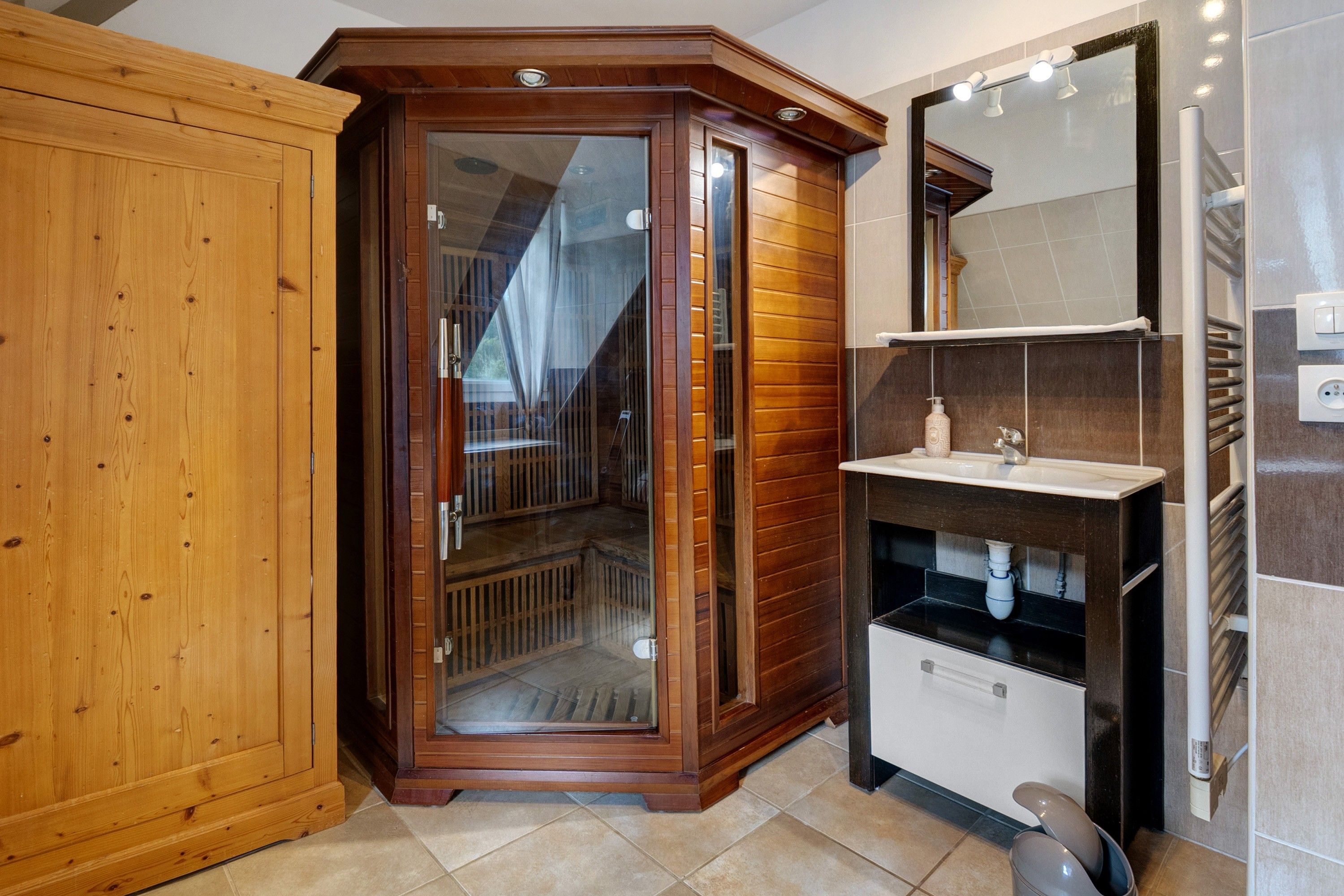 A corner room with a wooden wardrobe, a glass-enclosed cabinet, a sink with a mirror, and a radiator.