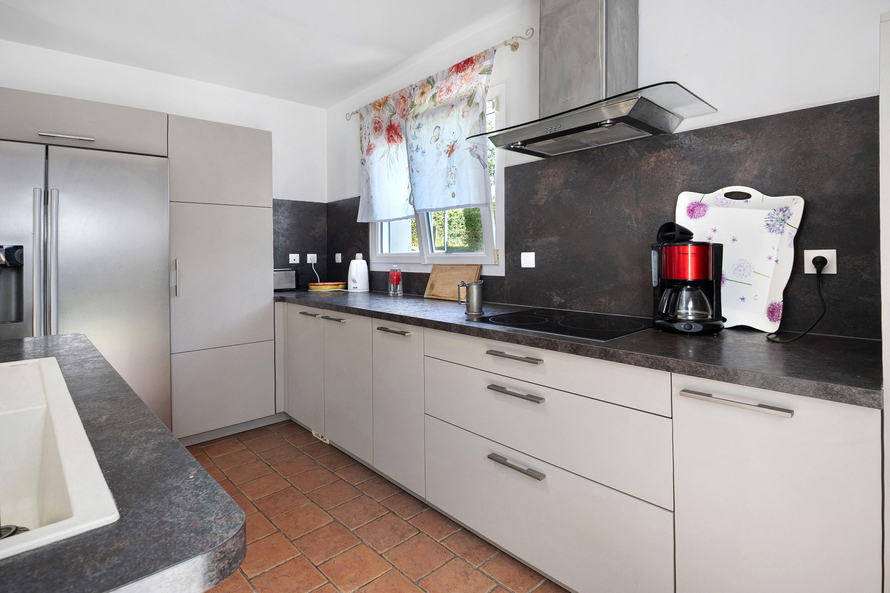 A modern kitchen with a built-in oven, a coffee maker, and a window with floral curtains.