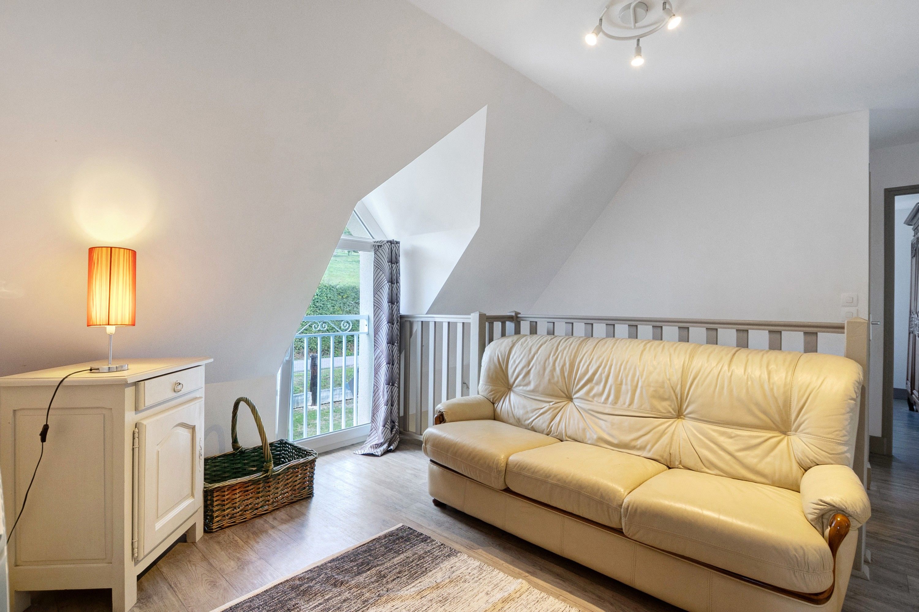 Ein Zimmer mit einem weißen Sofa, einer Lampe, einem Korbwannenstuhl und Blick auf Grünflächen.