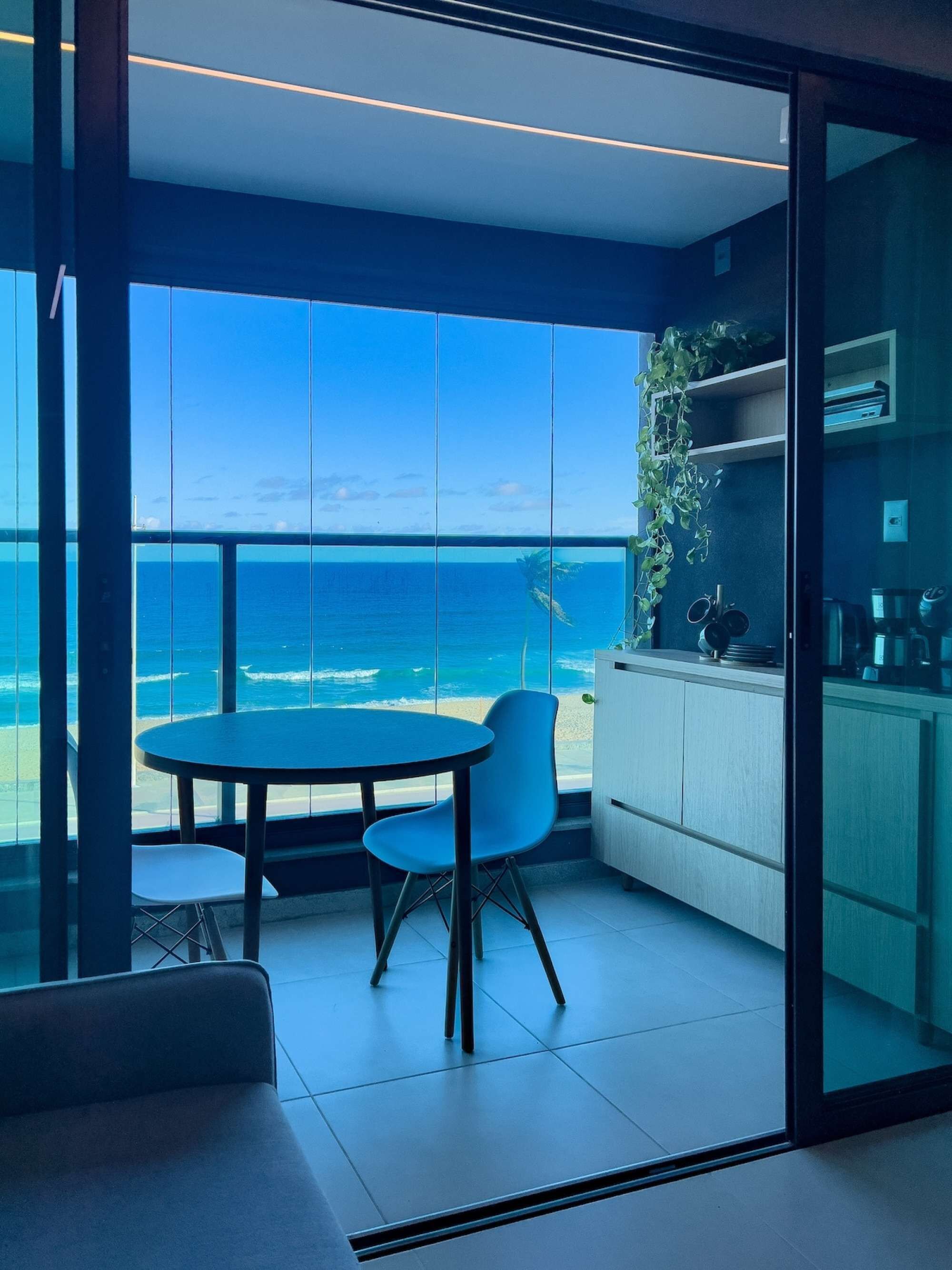 Uma varanda com vista para o oceano, uma mesa redonda, duas cadeiras e uma cozinha com armários e uma planta.