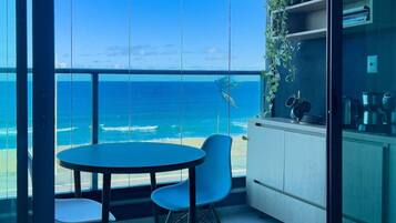 Uma varanda com vista para o oceano, uma mesa redonda, duas cadeiras e uma cozinha com armários e uma planta.