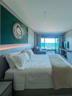 A modern hotel room with a large bed, a TV, and a balcony with a view of the sea.