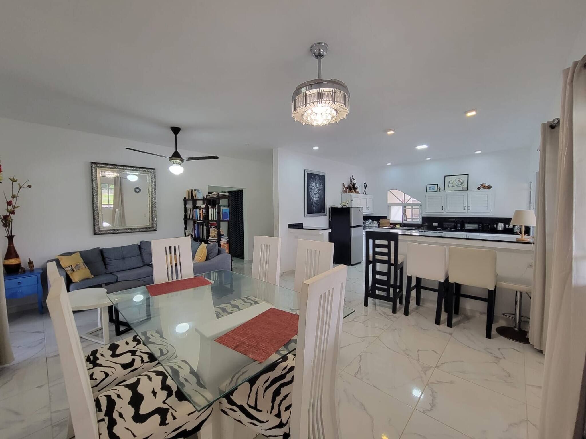 A modern living room with a glass dining table, a ceiling fan, and a kitchen area with white cabinets and a black countertop.
