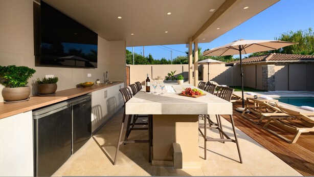 An outdoor kitchen area with a dining table, chairs, and a poolside setting. - Villa Idola Dassia Corfu (Dassia)