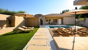 A poolside area with lounge chairs and umbrellas, a clear sky, and a view of a house in the background. - Villa Idola Dassia Corfu (Dassia)