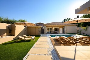 A poolside area with lounge chairs and umbrellas, a clear sky, and a view of a house in the background. - Villa Idola Dassia Corfu (Dassia)
