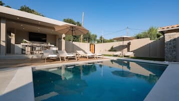 Une piscine entourée de chaises longues et de parasols, ainsi que d’un patio aménagé avec un téléviseur et du mobilier d’extérieur.