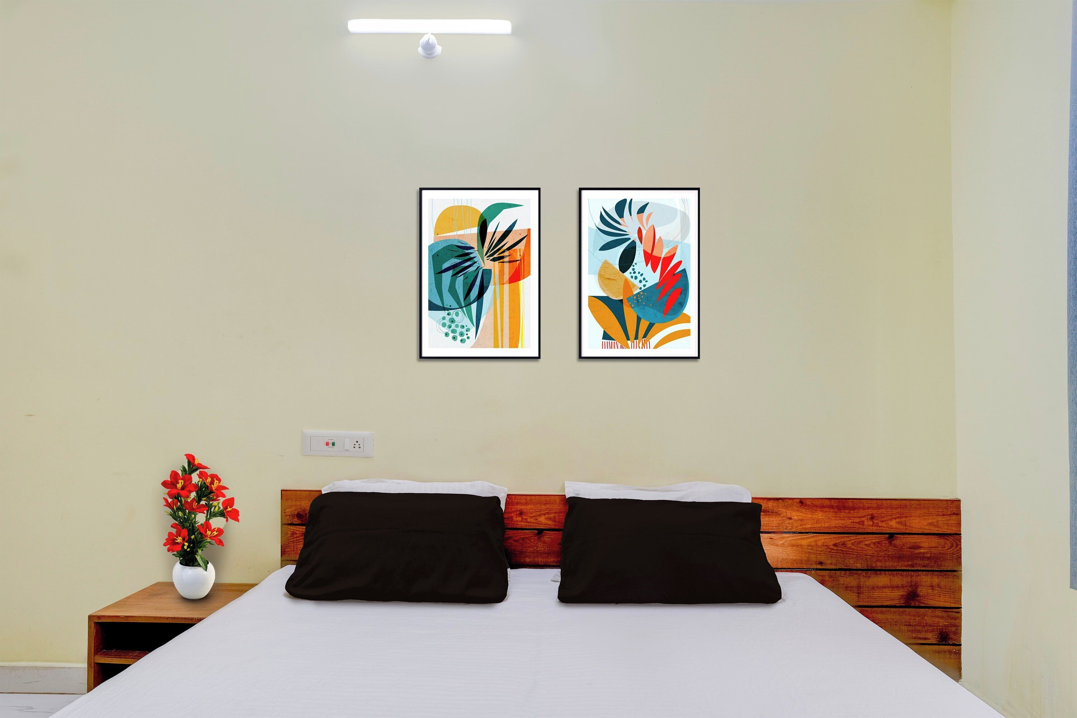 Habitación de hotel con cama, dos almohadas, mesita de noche con un florero y dos cuadros abstractos en la pared.