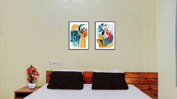 Habitación de hotel con cama, dos almohadas, mesita de noche con un florero y dos cuadros abstractos en la pared.